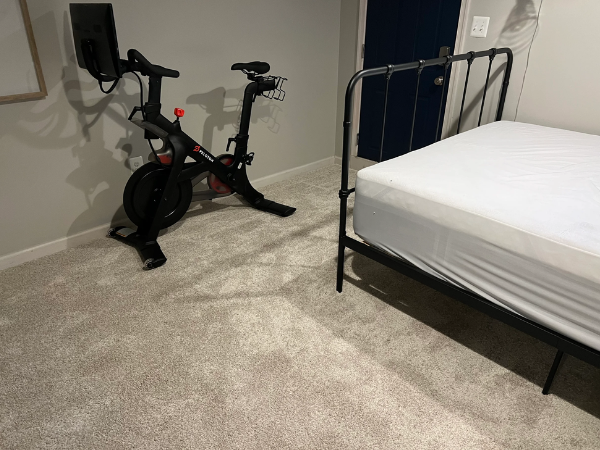 Grey carpet installation in guest room with indoor stationary bike and a black metal bed with mattress.